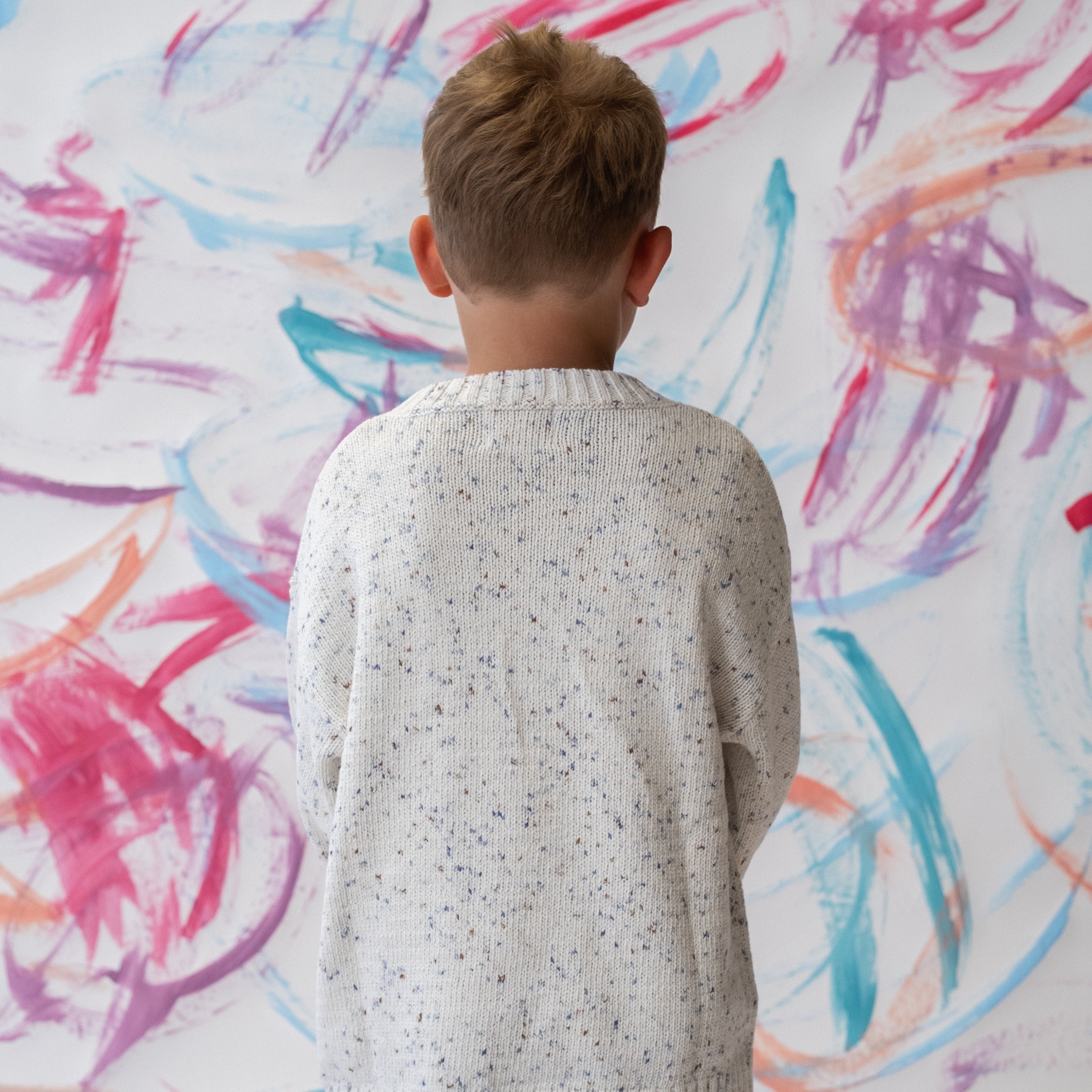 Child wearing a funfetti pullover sweater in surf standing in front of a colorful abstract wall.