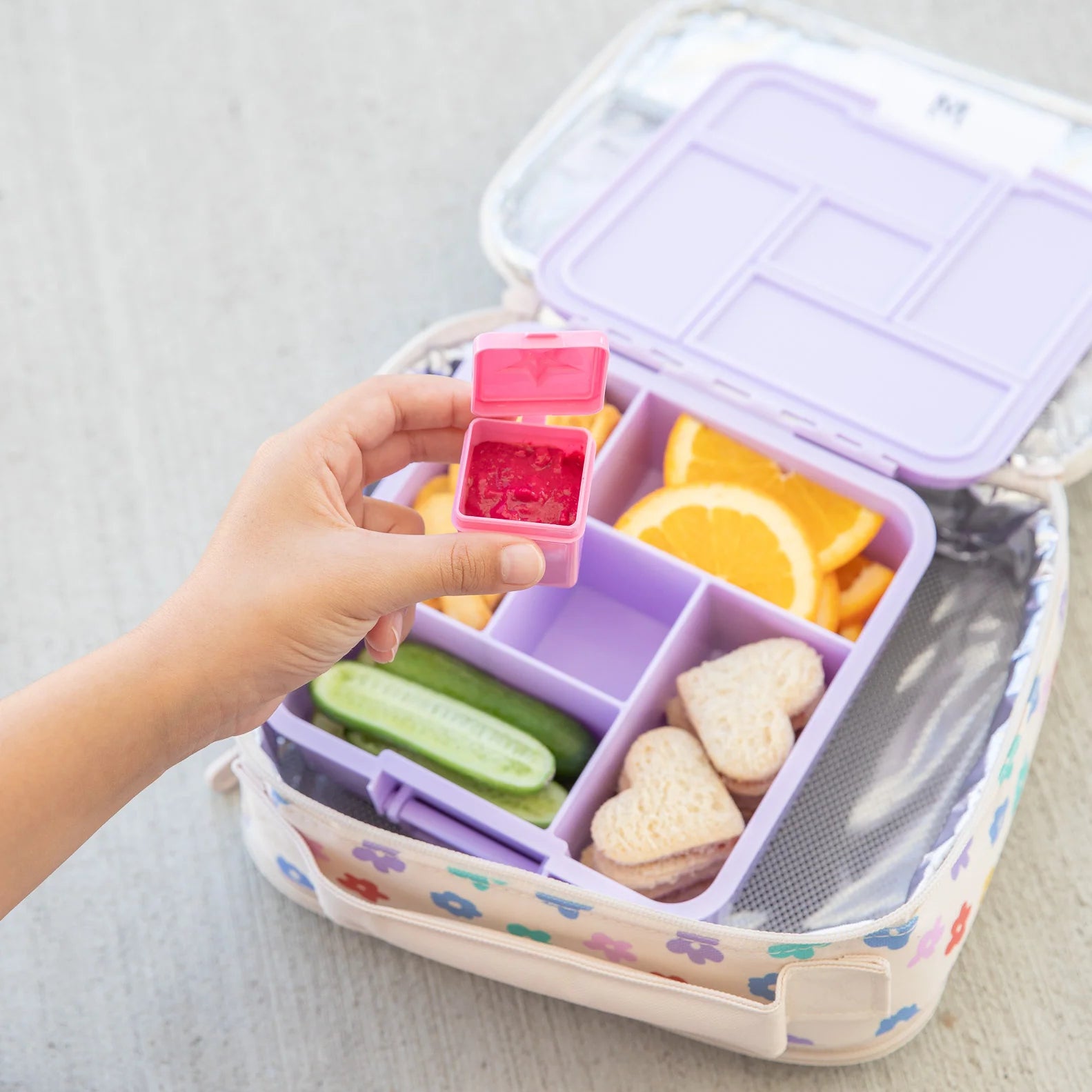 Bento box with compartments filled with snacks, including a pink container held by a hand.