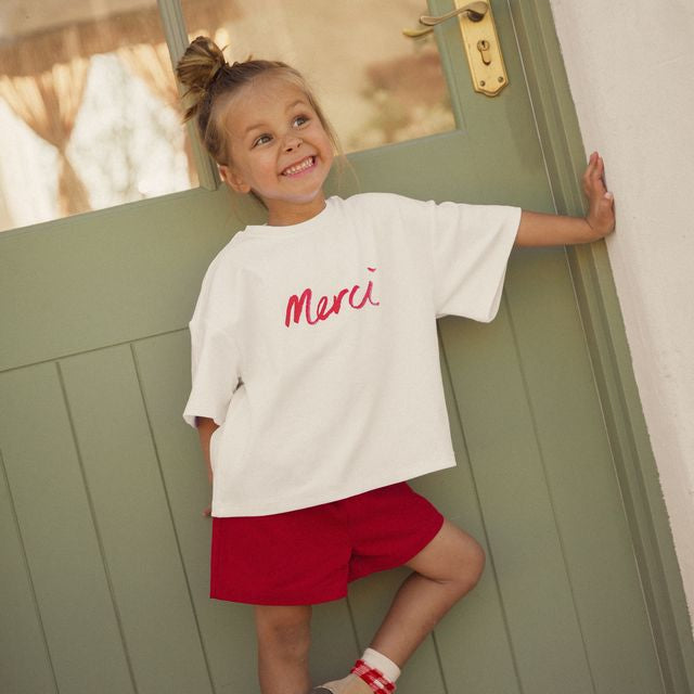 Child wearing a white shirt with 'Merci' in red and red shorts standing in front of a green door. Cinnamon Baby Merci collection