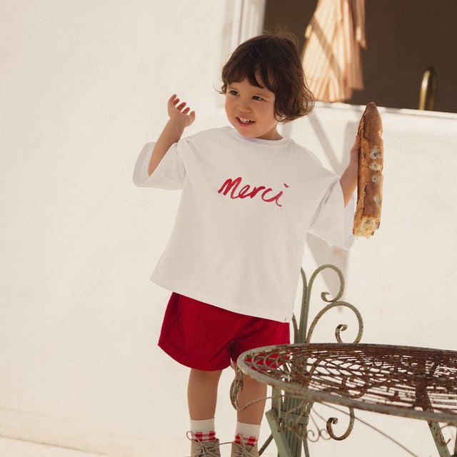 Child wearing a white shirt with 'Merci' in red text, standing on a metal chair outdoors. Merci collection by White Fox & Co