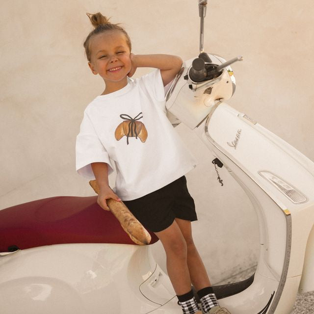 Child sitting on a scooter wearing a white t-shirt with a crossiant design by Cinnamon Baby