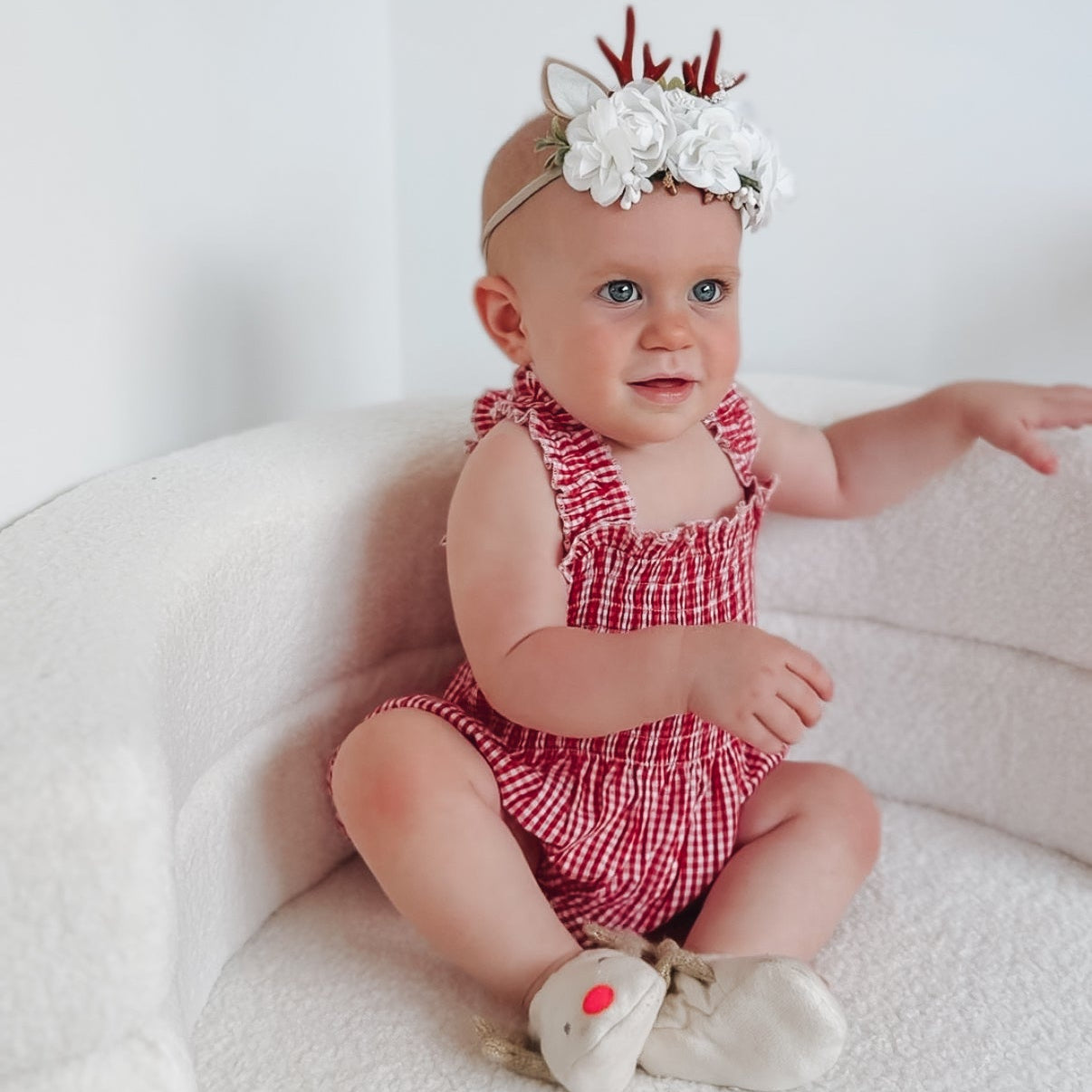 Baby in a red checkered romper with floral headband sitting on a white couch.