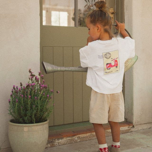 Cinnamon Baby Tee, postage stamp print, White shirt with a graphic of a red scooter on a beige background, resembling a vintage postcard, on a white background. White Fox & Co stockist. 
