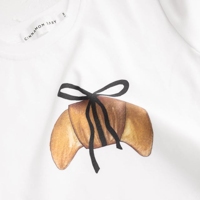 Child sitting on a scooter wearing a white t-shirt with a crossiant design by Cinnamon Baby