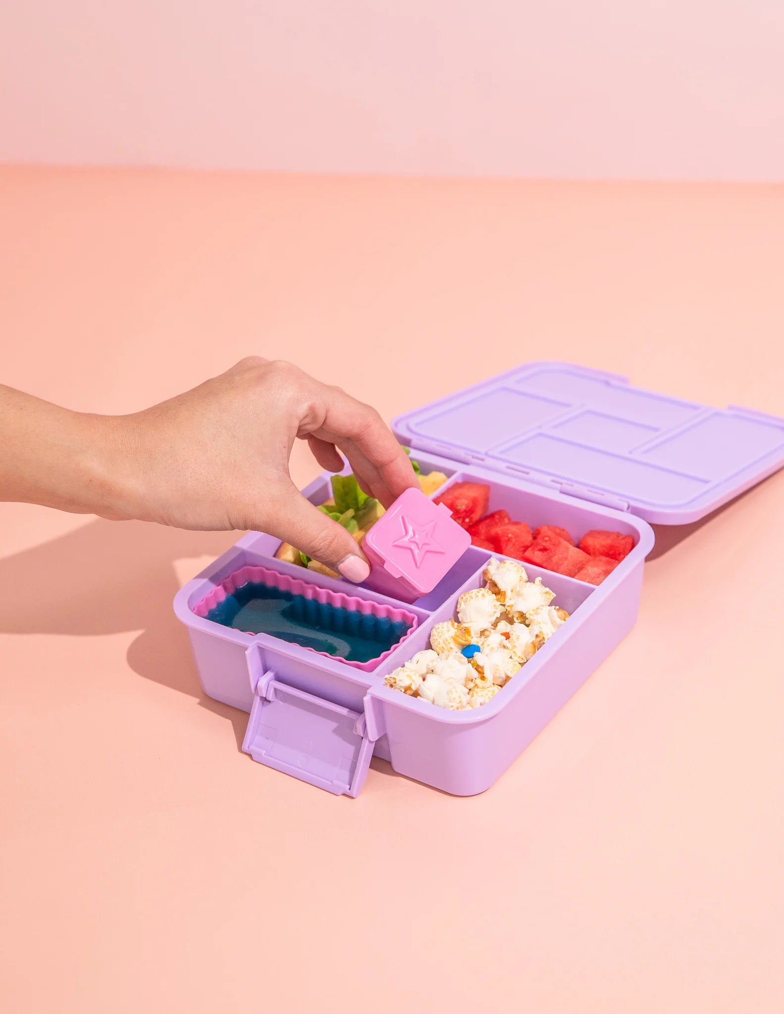 Purple bento box with compartments filled with snacks on a pink background