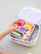 Bento box with compartments filled with snacks, including a pink container held by a hand.