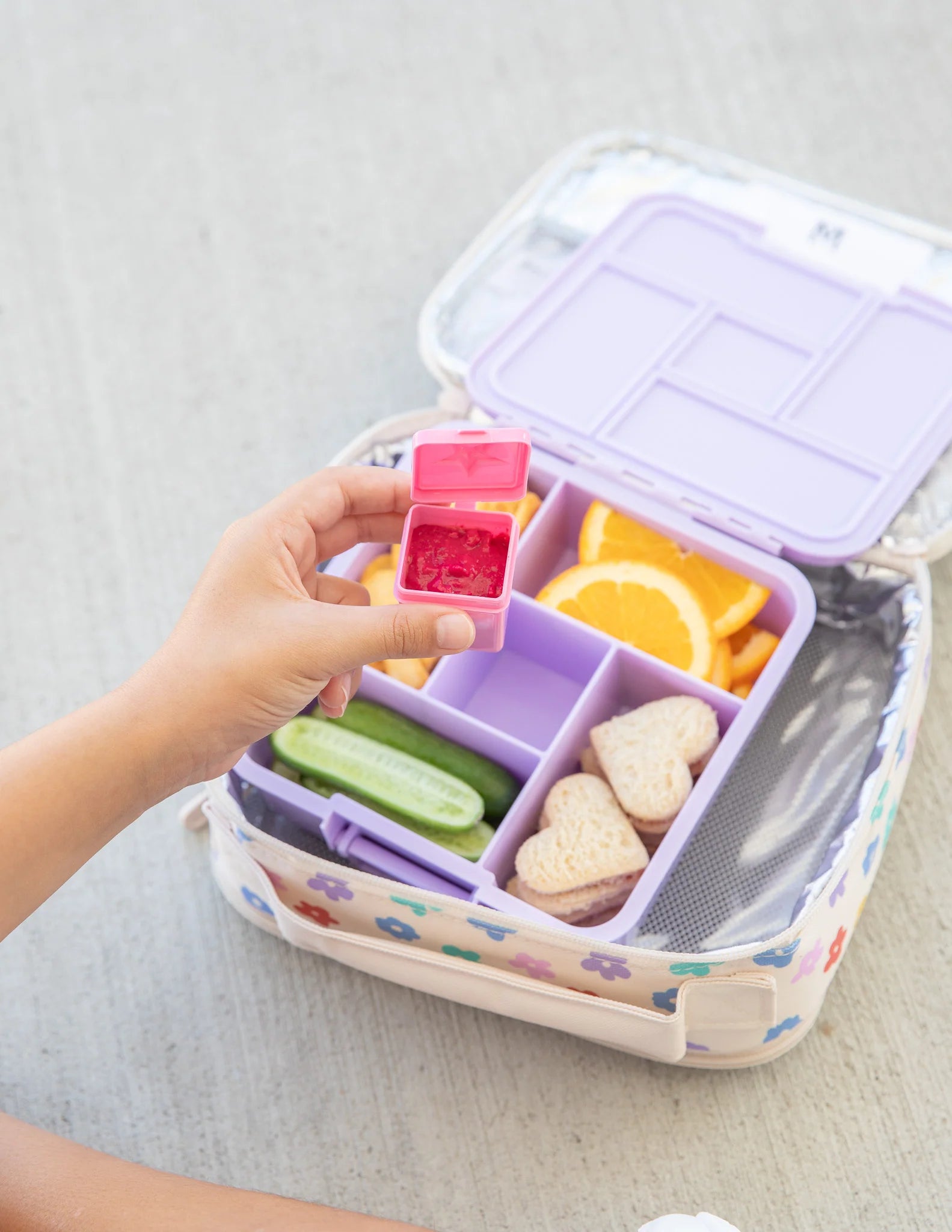 Bento box with compartments filled with snacks, including a pink container held by a hand.