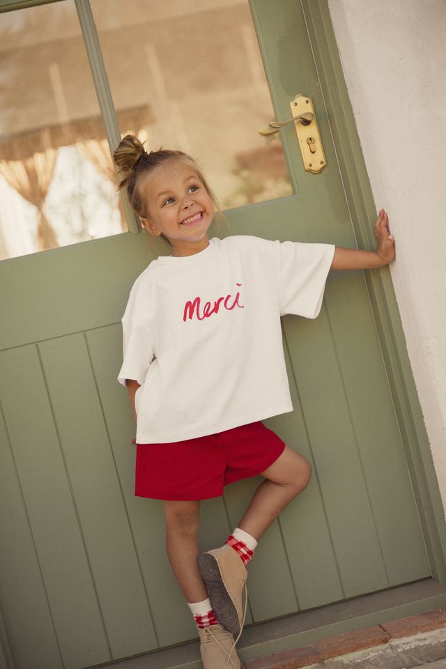Child wearing a white shirt with 'Merci' in red and red shorts standing in front of a green door. Cinnamon Baby Merci collection