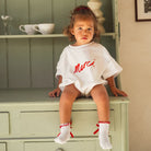 Child sitting on a green cabinet wearing a white romper with red text saying Merci by Cinnamon Baby