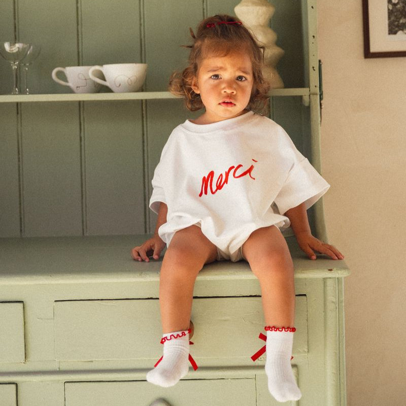 Child sitting on a green cabinet wearing a white romper with red text saying Merci by Cinnamon Baby