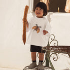 Child holding a baguette outdoors with wearing white tee with croissant print by Cinnamon Baby.
