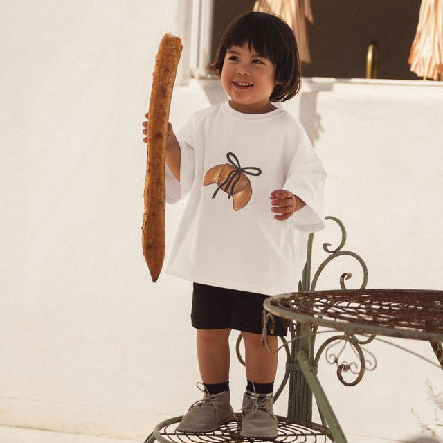 Child holding a baguette outdoors with wearing white tee with croissant print by Cinnamon Baby.