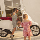 Two children in matching checkered outfits standing next to a white scooter on a patio. Merci Cinnamon Baby collection